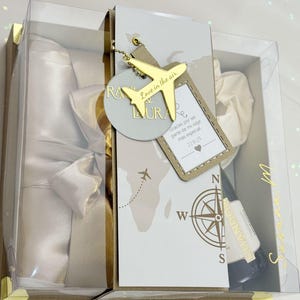 Puede incluir: Caja de regalo transparente con detalles dorados. Contiene una bata de satén blanco, una botella pequeña y un charm de avión dorado con la inscripción "Love in the air". Tema de viaje.