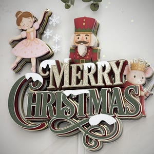 Op de afbeelding: Een gelaagde kerstversiering met een ballerina, notenkraker en muizenkoning. De woorden "MERRY CHRISTMAS" zijn in goud en groen glitter. De ballerina draagt een roze jurk en de notenkraker heeft een rood uniform.