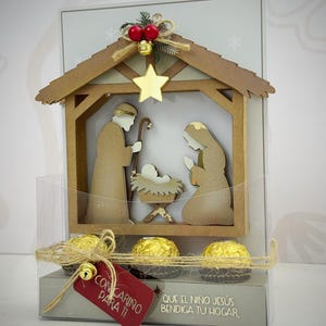 Könnte beinhalten: Ein weihnachtliches Geschenkset mit einer Holz-Krippe, einem goldenen Stern und roten Beeren. Das Set enthält drei goldverpackte Pralinen und ein rotes Etikett mit "Con cariño para ti". Der Text "QUE EL NIÑO JESÚS BENDIGA TU HOGAR" ist ebenfalls sichtbar.