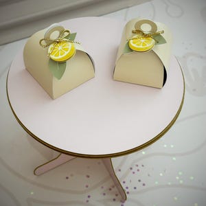 May include: Two cream-colored arched favor boxes, each topped with a yellow lemon slice cutout, a green leaf accent, and a thin gold metallic cord tied in a small bow. The boxes feature a circular handle cutout at the top of the arched structure.
