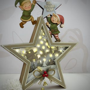 May include: A star-shaped Christmas decoration with a layered design. The interior features a silhouette of Santa's sleigh and reindeer, illuminated by small lights. Two elf figures are positioned on the star, and the text "ELIA POGGIOLI ROSAS" is visible.