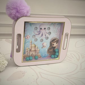 Puede incluir: Un juguete de madera rosa con temática de sirenas. El juguete presenta una imagen de un pulpo, un castillo y una sirena. Tiene un pompón morado en la parte superior y cuentas azules en la parte inferior. El juguete tiene un asa a cada lado.