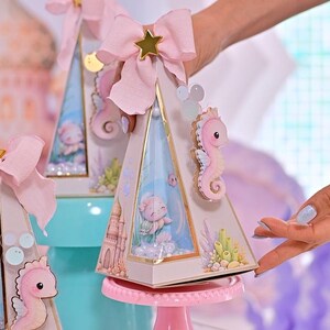K&ouml;nnte beinhalten: Dekorative pyramidenf&ouml;rmige Schachteln in Rosa und Gold mit Meerjungfrauen-Motiv. Jede Schachtel hat ein rosa Seepferdchen, eine rosa Schleife mit einem goldenen Stern und eine Meerjungfrauenszene im Inneren. Die Schachteln werden auf einem rosa Kuchenst&auml;nder pr&auml;sentiert.