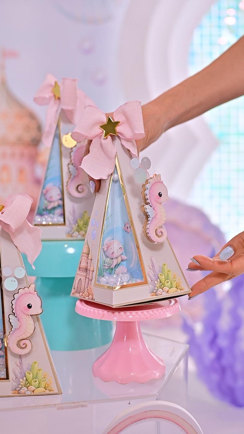 K&ouml;nnte beinhalten: Dekorative pyramidenf&ouml;rmige Schachteln in Rosa und Gold mit Meerjungfrauen-Motiv. Jede Schachtel hat ein rosa Seepferdchen, eine rosa Schleife mit einem goldenen Stern und eine Meerjungfrauenszene im Inneren. Die Schachteln werden auf einem rosa Kuchenst&auml;nder pr&auml;sentiert.