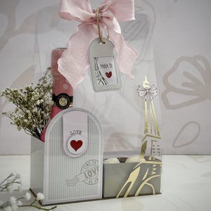 Op de afbeelding: Een cadeaupakket met een roze strik, een fles roze champagne en een decoratieve Eiffeltoren doos. De mand bevat een label met de tekst "Para Ti" en een hart. De geschenkset is versierd met witte bloemen.