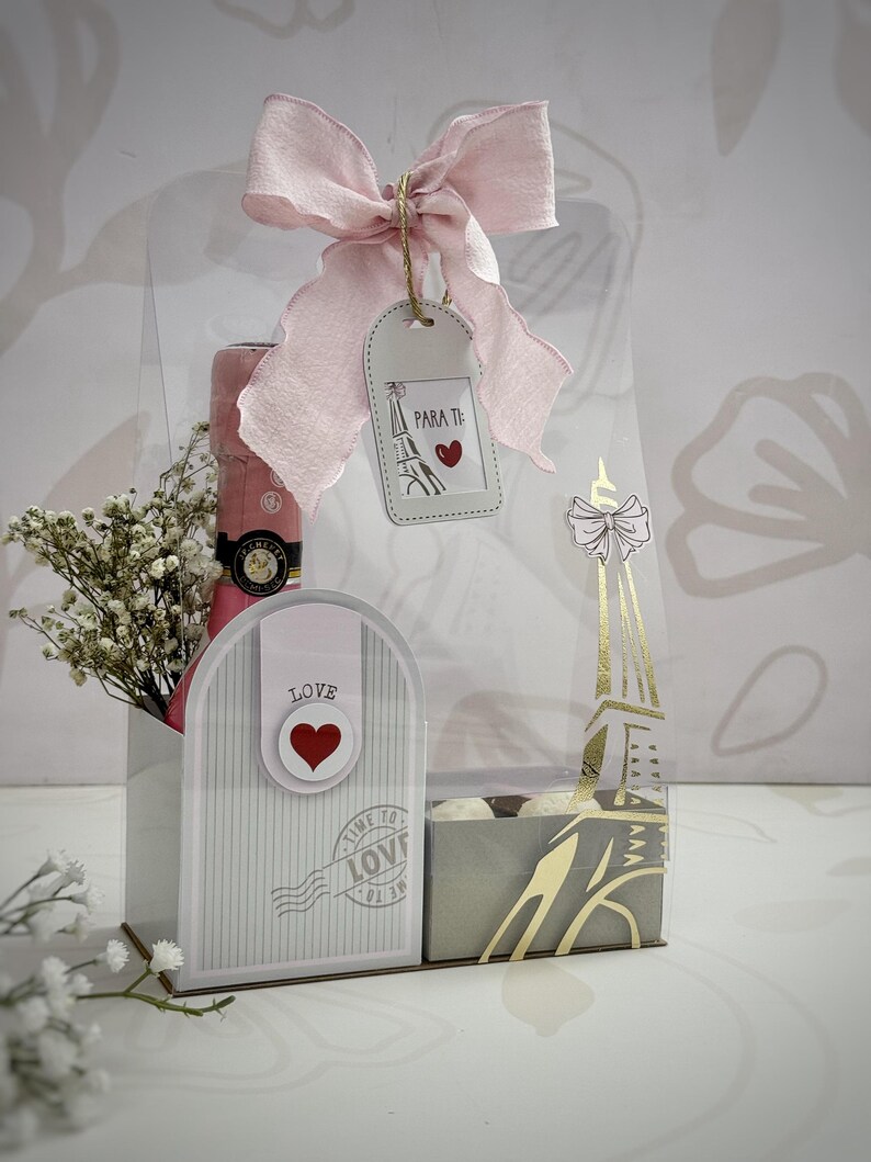 Op de afbeelding: Een cadeaupakket met een roze strik, een fles roze champagne en een decoratieve Eiffeltoren doos. De mand bevat een label met de tekst "Para Ti" en een hart. De geschenkset is versierd met witte bloemen.
