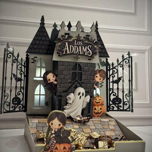 Op de afbeelding: Een decoratieve display met een miniatuurkasteel met de tekst Los Addams, zwarte hekken in ijzerstijl en cartoonachtige figuren, waaronder een meisje met vlechten, een geest en pompoenen. De structuur heeft een stenen ondergrond met snoepjes.