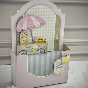 May include: A decorative paper card holder featuring a light pink and blue striped design. The front displays a layered cutout of a lemonade stand with a pink and white umbrella, a jar of lemons, and the text limonada, Disfruta tu día, and Dulce limón.