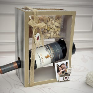 Puede incluir: Una caja de regalo con una botella de vino, pistachos y una foto. La caja es a rayas con un marco dorado y atada con hilo. La botella de vino está etiquetada como "Morande Pionero". También se incluye una foto pequeña y una decoración en forma de cámara.