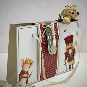 May include: A decorative gift bag with a red and white diamond pattern, featuring illustrations of a mouse king and a nutcracker. The bag has a gold ribbon handle and a small crocheted teddy bear keychain.