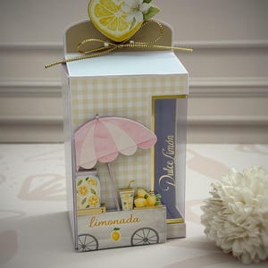May include: A rectangular paper box with a light yellow gingham pattern, featuring a heart-shaped lemon topper with a gold string bow. The front displays a pink and white striped umbrella, a lemonade stand graphic, and the text Dulce Limón.