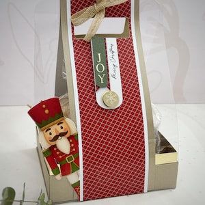 May include: A Christmas-themed gift bag with a red and white patterned design, a gold trim, and a clear plastic front. The bag is adorned with a beige bow and a tag that reads "JOY" and "Merry Christmas". A nutcracker figure peeks out from the side.