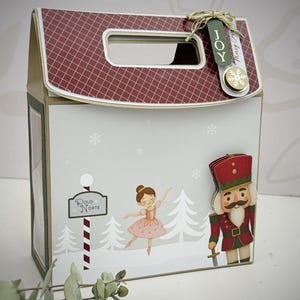 May include: A decorative Christmas gift box with a red and white roof design. The box features illustrations of a ballerina, a nutcracker, and a sign that says "Polo Norte". The box is decorated with snowflakes and the word "JOY".