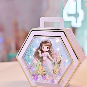 Op de afbeelding: Een zeshoekige, lichtroze decoratieve doos met een handvat, met een zeemeermin illustratie binnenin. De doos heeft gouden randen en bevat kleine lampjes, waardoor een verlichte display ontstaat. Het woord "Zoe" is zichtbaar op de achtergrond.