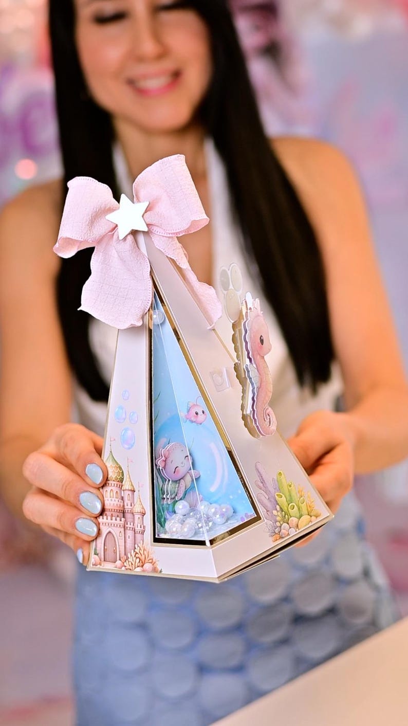 K&ouml;nnte beinhalten: Eine wei&szlig;e, dreieckige Geschenkbox mit einer rosa Schleife und einem Stern. Die Box zeigt ein Meerjungfrauen-Thema mit Illustrationen eines Schlosses, eines Seepferdchens und einer Unterwasserszene. Die Box wird von einer Person gehalten.