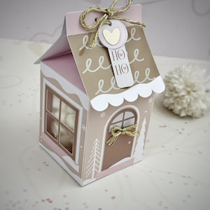 Puede incluir: Una caja de regalo decorativa con forma de casa, en tonos rosa y beige, con ventana y puerta. El tejado es rosa, y la caja está adornada con un lazo dorado y una etiqueta que dice "HO HO". La caja está decorada con detalles blancos y un corazón dorado.