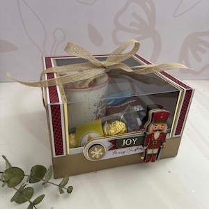 Pode incluir: Uma caixa de presente festiva com tampa transparente, amarrada com um laço de serapilheira. A caixa contém uma caneca, chocolates e outras guloseimas. Uma figura decorativa de quebra-nozes e uma faixa que diz "JOY Merry Christmas" estão anexadas.