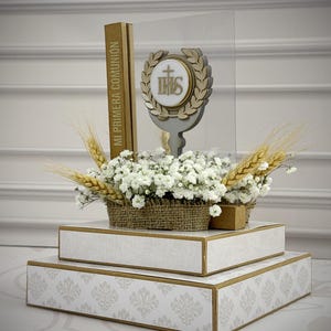以下が含まれることがあります： 段になった白と金の装飾ディスプレイ。花のアレンジメントと木製のエンブレムが特徴です。エンブレムには、十字架と、月桂樹の冠の中の文字IHSが描かれています。「MI PRIMERA COMUNION」の文字が見えます。