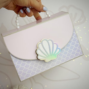 Puede incluir: Un bolso rosa claro con asa de perlas y cierre de concha. El bolso tiene un patrón de escamas de sirena en la mitad inferior. El cierre de concha tiene un contorno dorado y detalles iridiscentes.