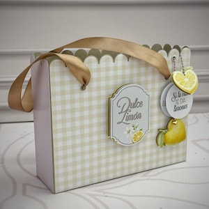 Puede incluir: Una bolsa de regalo rectangular con estampado de cuadros vichy en beige claro y blanco, borde superior festoneado y asas de cinta dorada. Una etiqueta central dice Dulce Limón con un gráfico de limón. Incluye etiquetas colgantes de limón.