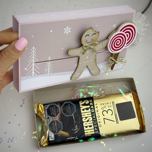 Könnte beinhalten: Eine rosa Geschenkbox mit Winterlandschaftsdesign, verziert mit einem Lebkuchenmann aus Holz und zwei rot-weißen Wirbellollis. Im Inneren ist eine Hershey's Special Dark Schokoladenriegel zu sehen.