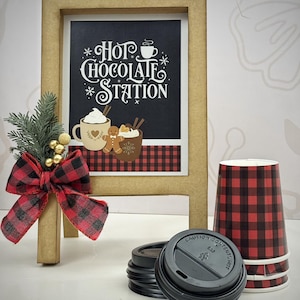 Puede incluir: Un letrero de madera con el texto "Hot Chocolate Station", un lazo a cuadros rojo y negro y vasos de papel. El letrero presenta ilustraciones de chocolate caliente y hombres de jengibre. También son visibles tapas negras y una pila de vasos a cuadros.