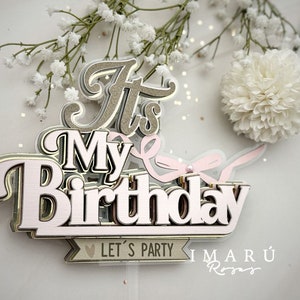 May include: It's My Birthday cake topper with pink and gold lettering and a pink ribbon. The topper has the text "Let's Party" on a banner and the text "Imaru Roses" below.