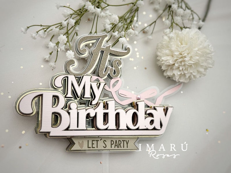 May include: It's My Birthday cake topper with pink and gold lettering and a pink ribbon. The topper has the text "Let's Party" on a banner and the text "Imaru Roses" below.