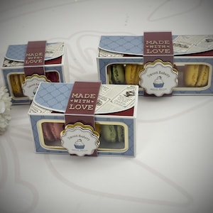May include: Three rectangular cardboard boxes containing colorful macarons, each featuring a blue and white lattice pattern, a maroon band labeled MADE WITH LOVE, and a scalloped tag with the text Sweet Bakery and a cupcake illustration.