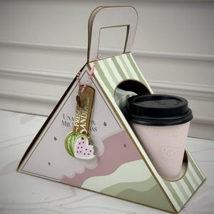 Puede incluir: Un portavasos triangular de cartón con asa, con rayas verdes y un patrón de sandía rosa. Contiene un vaso de papel rosa claro con tapa negra. Incluye una etiqueta rectangular con el texto "UNA MIL SANDIA" y un dije de sandía colgando.