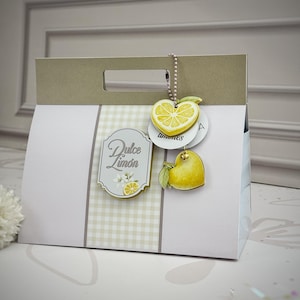 Könnte beinhalten: Eine weiße Geschenktüte aus Papier mit beigem Griff und einem zentralen vertikalen Feld mit hellbeigem Vichy-Muster. Ein Etikett trägt die Aufschrift Dulce Limón mit Zitronengrafik. Zwei zitronenförmige Anhänger hängen an einer silbernen Kette.