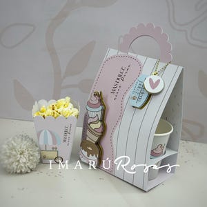 Peut inclure: Un sac cadeau décoratif avec une poignée rose et un motif à rayures, avec un motif de cupcake et de beignet. Un récipient à pop-corn et une fleur décorative sont également présents. Le sac porte l'inscription "MAS DULCE menos Drama".