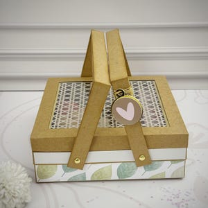 Puede incluir: Una caja de regalo decorativa con asa, con un patrón tejido en la parte superior y un diseño de hojas en la parte inferior. La caja es de cartón con un asa dorada y un dije en forma de corazón.
