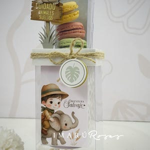 May include: A clear box with a jungle theme, containing macarons and decorative elements. The box features a sign that reads "Cuidado Animales Sueltos" and an illustration of a boy riding an elephant. The macarons are yellow, pink, and green.