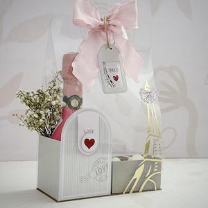 Pode incluir: Uma caixa de presente com um laço rosa, uma garrafa de champanhe rosa e um pequeno buquê de flores brancas. A caixa tem um design de caixa de correio com a palavra "LOVE" e um coração. Uma etiqueta diz "PARA TI" com uma ilustração da Torre Eiffel.