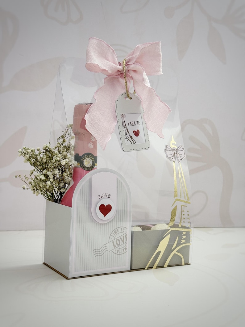 Op de afbeelding: Een geschenkdoos met een roze strik, een fles roze champagne en een klein boeket witte bloemen. De doos heeft een brievenbusontwerp met het woord "LOVE" en een hart. Een label zegt "PARA TI" met een illustratie van de Eiffeltoren.