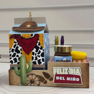 Può includere: Una scatola regalo in cartone con un design a tema cowboy, caratterizzata da un cappello marrone, una bandana rossa e un gilet con stampa a macchie di mucca. La base include un cactus verde, una ruota di carro, il nome Andy e un cartello.