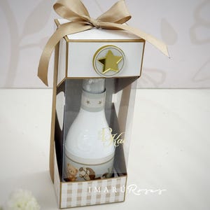 May include: A white baby bottle with a teddy bear and horse design, presented in a gift box with a gold star and bow. The box has a clear front and a checkered pattern at the base.