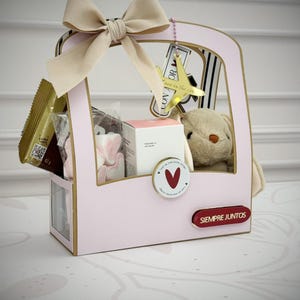 Op de afbeelding: Een roze geschenkdoos met een beige strik, met een pluchen konijn, een kleine witte doos en verpakte lekkernijen. Een gouden vliegtuigbedel hangt erin. De doos heeft de tekst "SIEMPRE JUNTOS" op de zijkant.