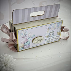 Puede incluir: Una caja de regalo decorativa con asa y lazo rosa. La caja presenta ilustraciones de dulces, incluyendo piruletas, macarons y un carrito de helados. El texto "Sweet Treats" es visible.