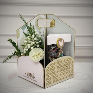 May include: A decorative gift box with a handle, featuring a floral arrangement and a compartment with candy. The box has a light pink front with the word "MAMÁ" and a heart symbol. The side has a woven pattern.
