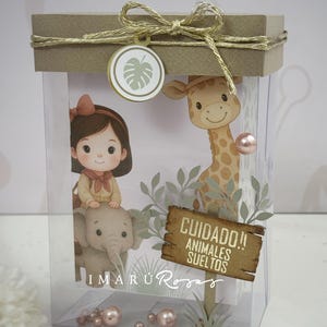 Puede incluir: Una caja transparente con tapa marrón atada con hilo dorado. En el interior, una ilustración de dibujos animados presenta a una niña sobre un elefante, una jirafa y un letrero que dice "Cuidado!! Animales Sueltos". La caja está decorada con perlas rosas y un dije en forma de hoja.