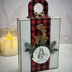 Puede incluir: Una caja de regalo blanca con una cinta y asa a cuadros rojos y negros. Un reno dorado y vegetación adornan el frente, con una etiqueta circular que dice "MERRY CHRISTMAS". Un lazo dorado adorna el asa.
