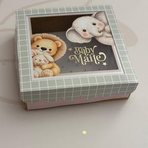 May include: A baby gift box in light grey and pink, decorated with illustrations of a lion, a bunny, and an elephant. The text "Baby Maite" is in gold lettering. The box has a grid pattern on the top and a scalloped edge.