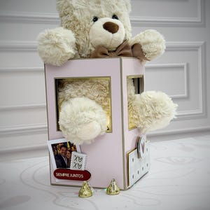 Puede incluir: Un oso de peluche crema con una pajarita marrón asoma de una caja rosa. La caja tiene detalles dorados y una foto con el texto "SIEMPRE JUNTOS". Dos bombones dorados están delante.