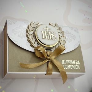 Könnte beinhalten: Eine dekorative Box mit goldenem Band und dem Text "MI PRIMERA COMUNIÓN". Die Box hat ein goldenes Emblem mit den Buchstaben IHS, einem Kreuz und einem Lorbeerkranz. Die Box hat ein weißes und goldenes Farbschema.
