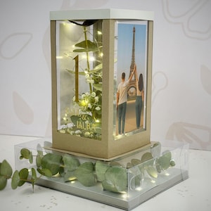 Puede incluir: Una caja decorativa con una foto de la Torre Eiffel y una pareja. La caja tiene un número 1 dorado y los nombres "& LAURA". Contiene vegetación y luces de hadas. La caja se encuentra sobre una bandeja transparente con más vegetación.