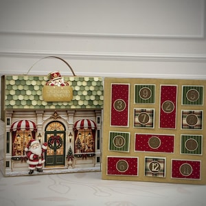 Puede incluir: Un calendario de adviento con forma de casa y asa, que muestra una figura de Santa Claus, una ilustración de escaparate y el texto "CALENDARIO DE ADVIENTO". Junto a él, un tablero cuadrado con doce puertas numeradas en tonos rojos y verdes.