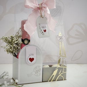 Op de afbeelding: Een cadeaupakket met een roze champagnefles, witte bloemen en een decoratieve doos met een roze strik. De set bevat een label met "PARA TI" met een hart en een illustratie van de Eiffeltoren. De doos heeft het woord "LOVE" en een hart.