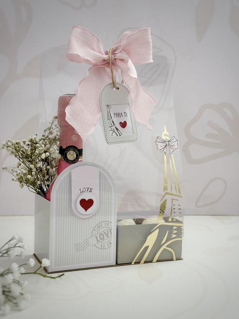 Op de afbeelding: Een cadeaupakket met een roze champagnefles, witte bloemen en een decoratieve doos met een roze strik. De set bevat een label met "PARA TI" met een hart en een illustratie van de Eiffeltoren. De doos heeft het woord "LOVE" en een hart.